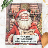 A square fabric napkin or tea towel featuring a festive Christmas illustration of Santa Claus in front of a fireplace, holding a sign that reads, 'if I'm good at being naughty which list do i go on?' The napkin is styled on a light marble surface with a wooden spoon, greenery, and a small Christmas tree nearby.