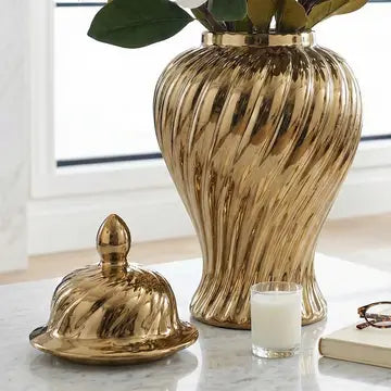 A shiny gold ribbed ceramic vase with a matching gold cloche lid, styled on a marble tabletop with a candle, book, and glasses in a bright room.