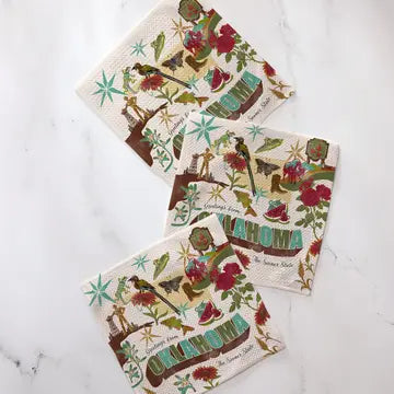 Three square cotton napkins with a vintage-style botanical and travel-themed print, arranged diagonally on a white marble surface.
