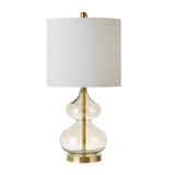 Table lamp with clear glass base, gold accents, and white shade on a white background