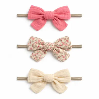 Three fabric hair bows on wooden sticks, arranged vertically on a white background. The top bow is solid pink, the middle bow has a small floral print on a beige background, and the bottom bow is solid cream.
