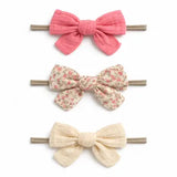 Three fabric hair bows on wooden sticks, arranged vertically on a white background. The top bow is solid pink, the middle bow has a small floral print on a beige background, and the bottom bow is solid cream.