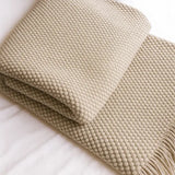A folded beige knit throw blanket with a textured waffle pattern and fringe, displayed on a white fabric background.