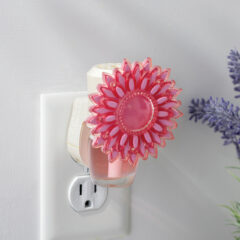 Pink flower-shaped night light attached to a wall outlet with a blurred plant in the background.