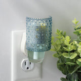 Plug-in night light with a blue textured glass cover on a white wall, next to a green plant.