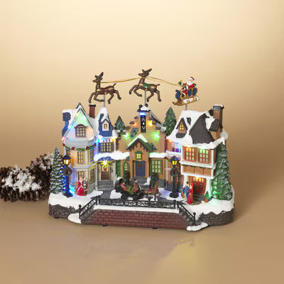 A festive miniature Christmas village figurine with snow-covered houses, glowing warm lights, a small bridge, evergreen trees, and Santa in a sleigh pulled by reindeer, displayed on a warm beige background.
