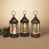 Three ornate black metal lanterns with hanging loops, decorated with festive Christmas scenes inside, including Santa, snowman, and Christmas tree figures. The lanterns are displayed on a beige background with a pinecone and red berries prop on the left.