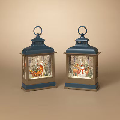 Two decorative lanterns with blue tops and bases, shown side by side on a warm beige background. Each lantern contains a small winter scene with a fox and a snowman in a snowy forest.