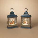 Two decorative lanterns with blue tops and bases, shown side by side on a warm beige background. Each lantern contains a small winter scene with a fox and a snowman in a snowy forest.