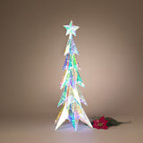 A decorative illuminated Christmas tree in a star-topped conical shape, covered in iridescent holographic material that shifts between green, blue, pink, and gold. It stands on a neutral warm brown background next to a red poinsettia stem.