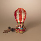 A festive hot air balloon Christmas ornament with a red and gold glittery balloon, Santa Claus and reindeer in the basket, and small cinnamon sticks and pinecones on a beige background.