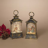 Two decorative lantern-style snow globe ornaments with hanging loops, shown on a beige studio background. One contains an angel figure and the other a nativity scene.
