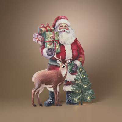 A festive Christmas-themed decorative figure or backdrop showing Santa Claus in a red suit holding wrapped gifts, standing beside a small lit Christmas tree and a brown deer, against a plain warm beige studio background.