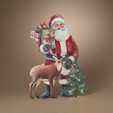A festive Christmas-themed decorative figure or backdrop showing Santa Claus in a red suit holding wrapped gifts, standing beside a small lit Christmas tree and a brown deer, against a plain warm beige studio background.