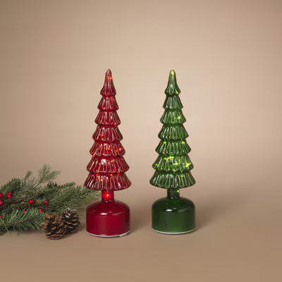 Two small Christmas tree-shaped glass ornaments in red and green, each mounted on a round base, displayed on a beige background with a pine branch and pinecone at the lower left.