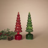 Two small Christmas tree-shaped glass ornaments in red and green, each mounted on a round base, displayed on a beige background with a pine branch and pinecone at the lower left.