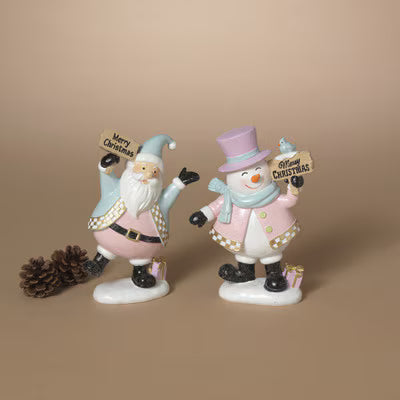 A pair of ceramic Christmas figurines, one of Santa Claus and one of a snowman, standing on a white surface against a solid beige background. Santa holds a sign reading 'Merry Christmas' and stands next to a pinecone.