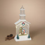 A white snow globe-style nativity scene housed in a church-shaped ornament, with a small figurine inside, a wreath on the front, and two small blue-green trees at the base. The ornament is photographed on a warm beige studio background with cinnamon sticks and a pinecone beside it.