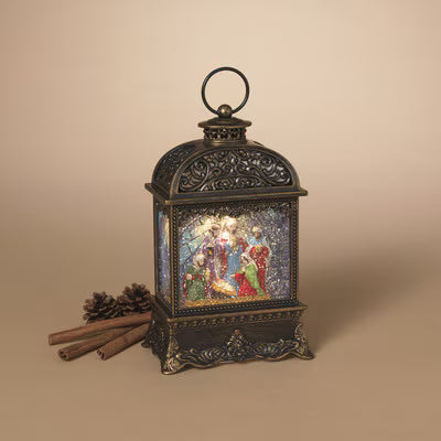 An ornate dark bronze lantern with a lit Christmas nativity scene on the front, styled on a beige background with cinnamon sticks and pinecones.