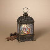 An ornate dark bronze lantern with a lit Christmas nativity scene on the front, styled on a beige background with cinnamon sticks and pinecones.