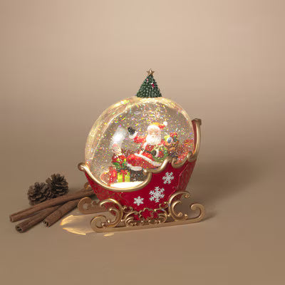 A festive Christmas snow globe in Santa's sleigh, featuring a red sleigh with gold scrollwork, a clear globe filled with glitter and Santa, a small green tree topper, and holiday decorations on a beige background.