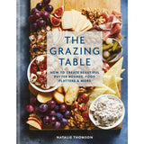 The Grazing Table: How to Create Beautiful Butter Boards, Food Platters & More by Natalie Thomson, featuring a beautifully styled gourmet charcuterie board with grapes, figs, crackers, cheese, salami, pickles, and fruit on a dark blue background.