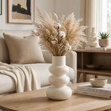 A cream-colored ceramic vase with a stacked, rounded design, filled with dried pampas grass, cotton bolls, and small wildflowers, set on a wooden coffee table in a modern, neutral-toned living room.