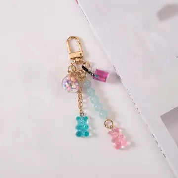 A gold-tone keychain bag charm featuring a lobster clasp, a clear bead filled with pastel glitter, a small pink bottle charm, light blue beads, a blue translucent bear, and a pink translucent bear on a white background.
