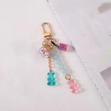 A gold-tone keychain bag charm featuring a lobster clasp, a clear bead filled with pastel glitter, a small pink bottle charm, light blue beads, a blue translucent bear, and a pink translucent bear on a white background.