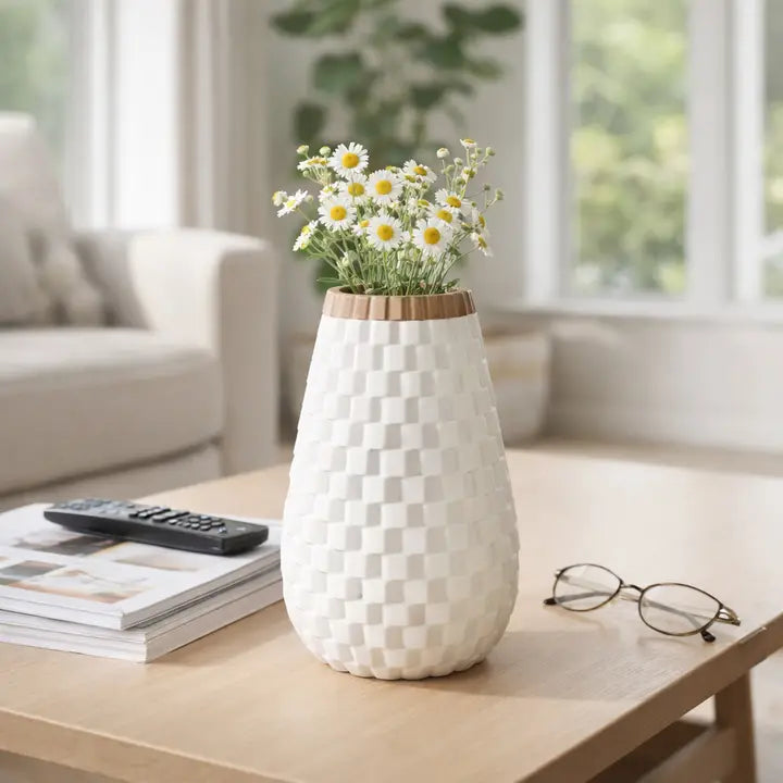 A white textured ceramic vase with a checkered pattern and a light wood rim, filled with white daisies, sitting on a light wood table in a bright, modern living room.