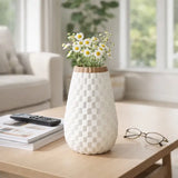 A white textured ceramic vase with a checkered pattern and a light wood rim, filled with white daisies, sitting on a light wood table in a bright, modern living room.