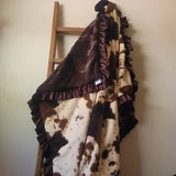 A plush throw blanket with a brown and cream cow-print pattern, dark brown ruffled trim, and a solid dark brown reverse side, draped over a wooden ladder against a plain wall.