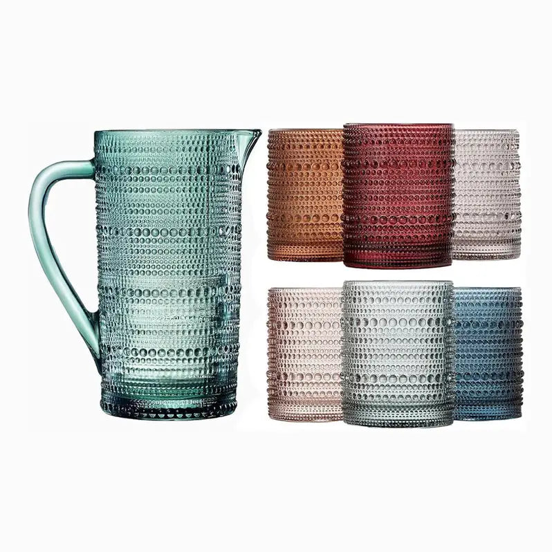 A textured glass pitcher in a translucent aqua-teal color stands on the left, with six matching textured tumblers in assorted colors arranged on the right against a plain white background.