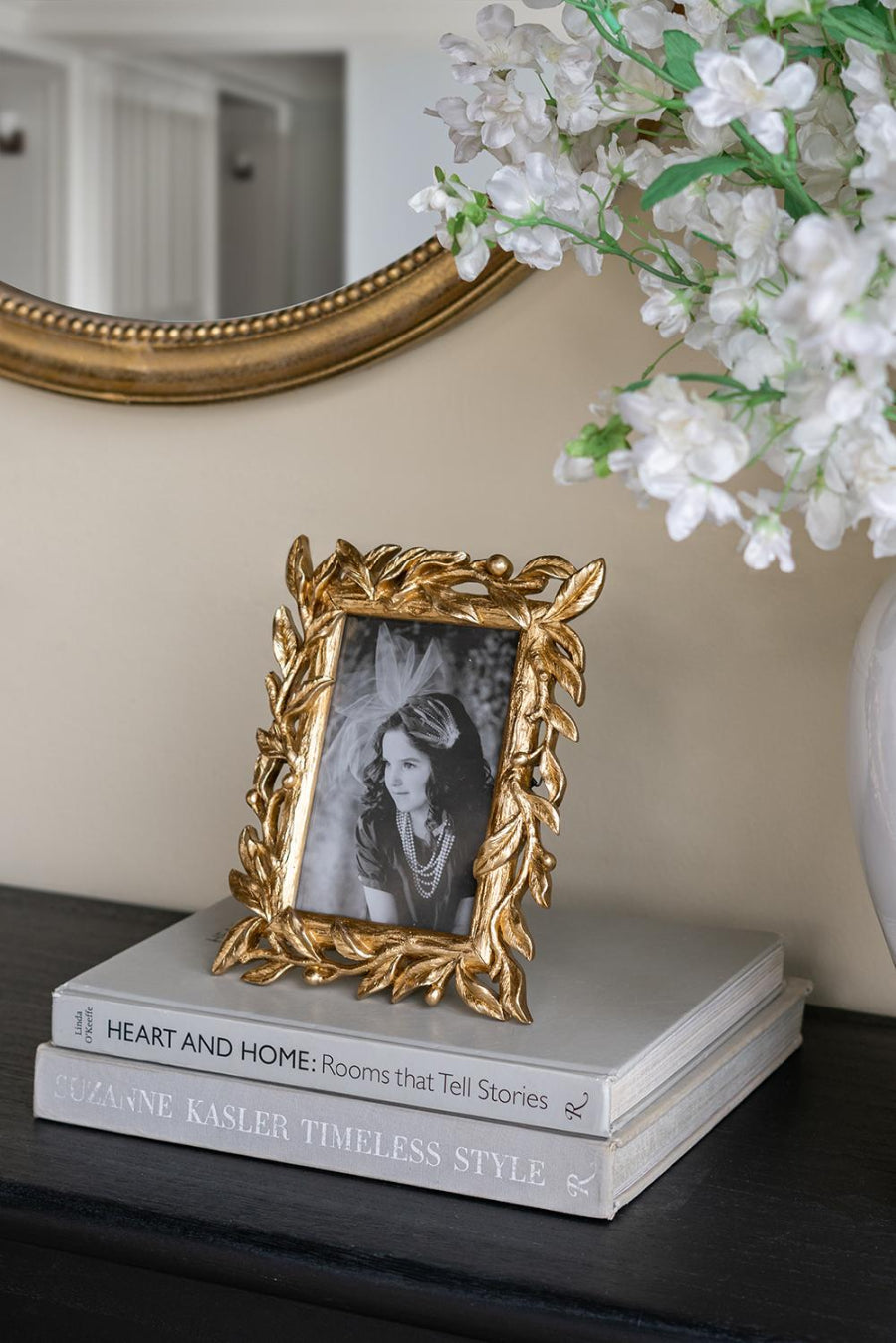 Decorative gold frame with a black and white photo on top of books, with a mirror and flowers in the background.