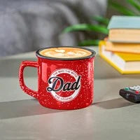A red ceramic mug with 'Dad' written on it, holding a beverage, placed on a surface with books and a remote control in the background.