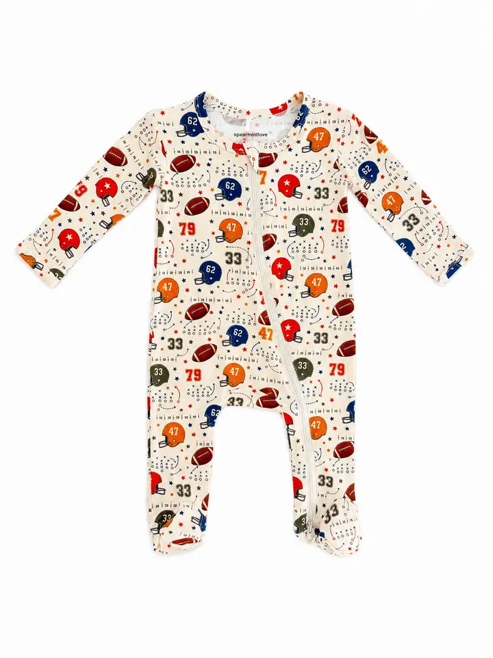 A baby long-sleeve footed sleeper with an all-over football-themed print on a cream background. The print features red, blue, and orange football helmets, footballs, numbers, and small stars.