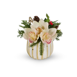 A decorative floral arrangement in a white pumpkin-shaped vase with vertical gold stripes. The arrangement features large cream-colored orchids with pinkish-red centers, green pine branches, a small pinecone, and red berries, set against a plain white background.