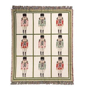A square woven throw blanket with fringe on all sides, featuring a grid of nine nutcracker illustrations on an off-white background with green borders.