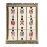 A square woven throw blanket with fringe on all sides, featuring a grid of nine nutcracker illustrations on an off-white background with green borders.