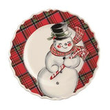 A round holiday plate with a scalloped edge, featuring a white snowman illustration wearing a black top hat and red plaid scarf and cane, set against a red and black tartan patterned background.