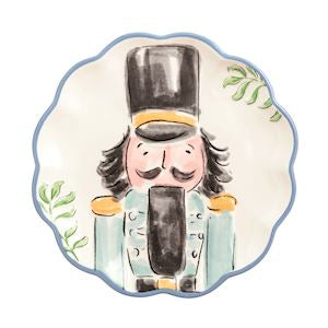 A scalloped ceramic plate featuring a hand-painted watercolor illustration of a nutcracker soldier with a black top hat, light blue coat, and green leaf accents on a white background.