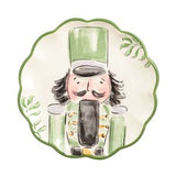 A scalloped ceramic plate featuring a hand-painted nutcracker illustration in green, black, and beige tones, with a green leaf motif in the upper right corner.