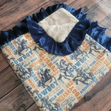 A folded western-themed fleece blanket or throw featuring a navy blue ruffled hood lining, a cream sherpa insert, and a printed body with cowboy silhouettes and text on a beige background, laid flat on a dark wood surface.