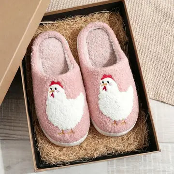 A pair of pink fuzzy slip-on house slippers with embroidered white chickens on the front, displayed inside a brown gift box filled with shredded paper.