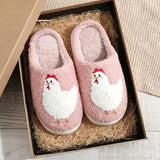 A pair of pink fuzzy slip-on house slippers with embroidered white chickens on the front, displayed inside a brown gift box filled with shredded paper.