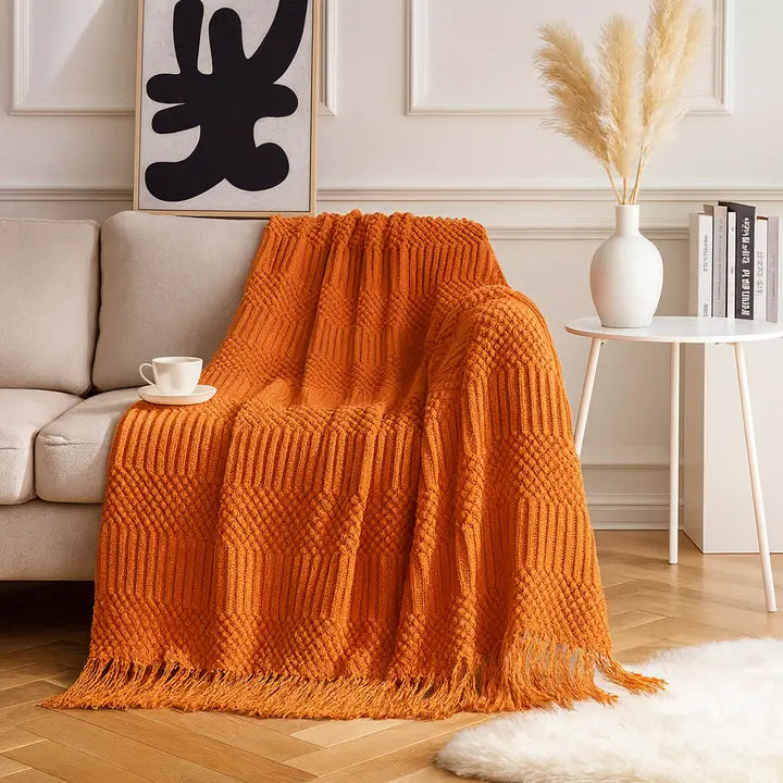 A bright orange chunky knit throw blanket with fringe is draped over a light beige sofa in a styled living room setting. The scene includes a white side table with a vase of pampas grass, books, a coffee cup, and a white rug on a wood floor.