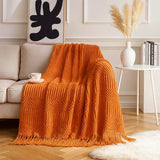 A bright orange chunky knit throw blanket with fringe is draped over a light beige sofa in a styled living room setting. The scene includes a white side table with a vase of pampas grass, books, a coffee cup, and a white rug on a wood floor.
