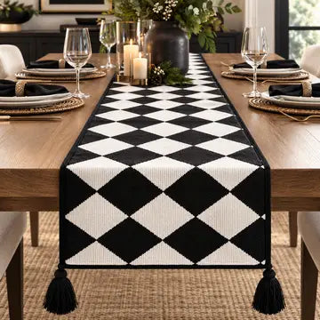 A black and white harlequin diamond-patterned table runner with black tassels, displayed on a wooden dining table set for four.