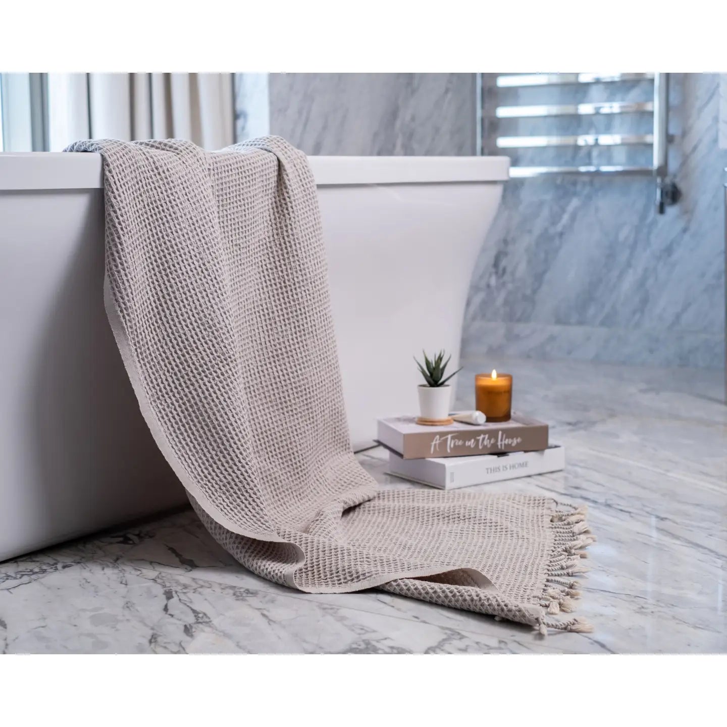 Beige textured blanket draped over a white bathtub with a candle and book on a marble surface.