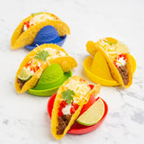 Four yellow hard taco shells arranged on colorful silicone taco holders on a white marble surface. The tacos are filled with ground meat, shredded cheese, sour cream, diced red pepper, and lime wedges.